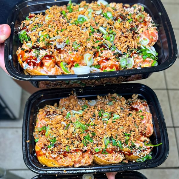 Two black containers filled with sushi, topped with fried onions, green onions, and sauce.