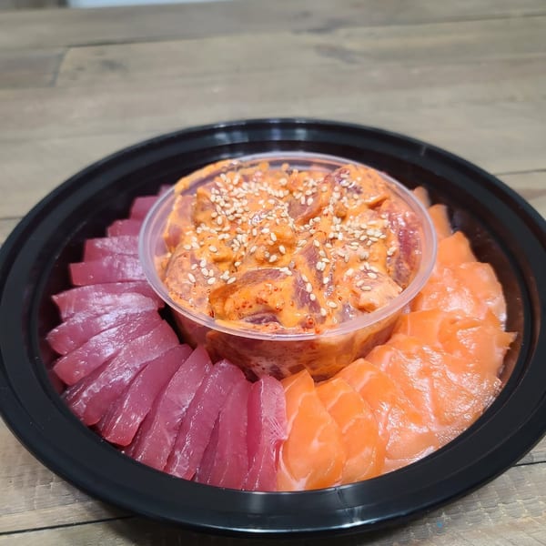 A black platter with a clear bowl of spicy tuna poke, surrounded by sliced tuna and salmon sashimi.