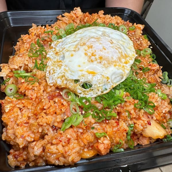 Kimchi fried rice topped with a fried egg, green onions, and sesame seeds in a black container.