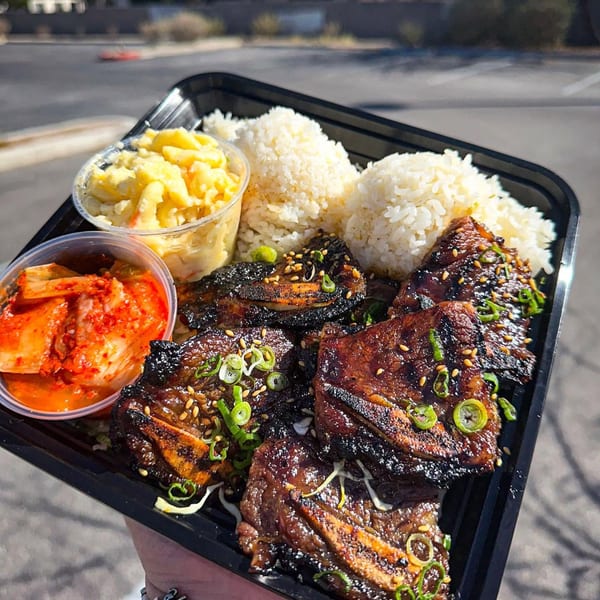 A takeout tray with grilled short ribs, white rice, macaroni salad, and kimchi, held outdoors.
