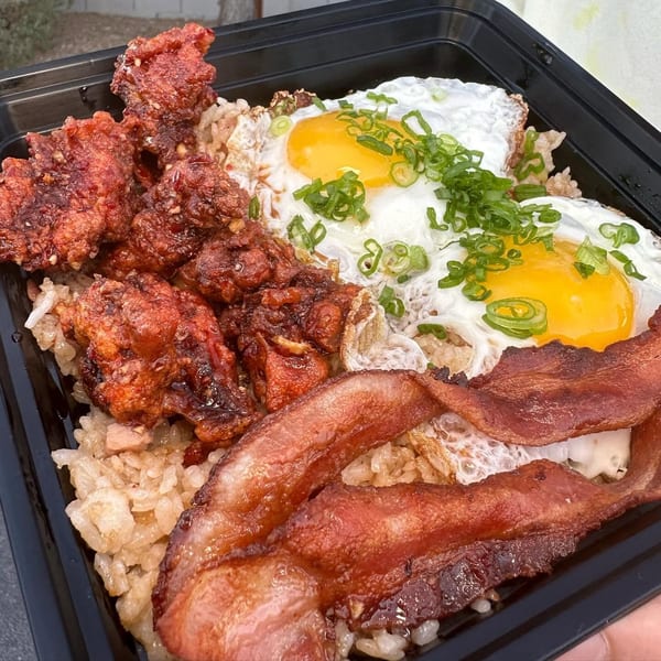 Fried chicken, sunny-side-up eggs with green onions, and bacon over rice in a black container.